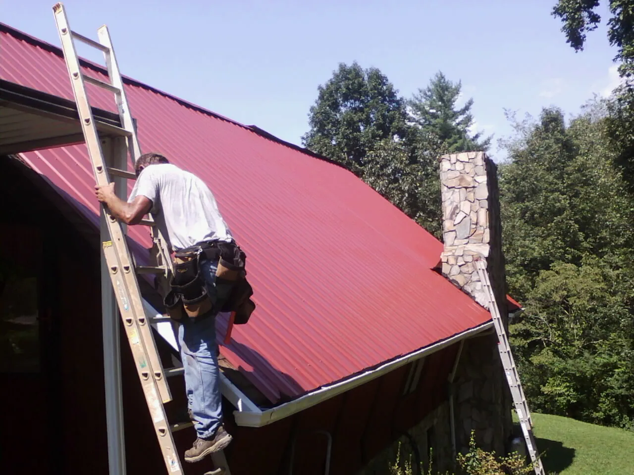 Roofer on a ladder beside a red metal roof and stone chimney — Soffit & Fascia Repair by Roofing Brothers in Doyle
